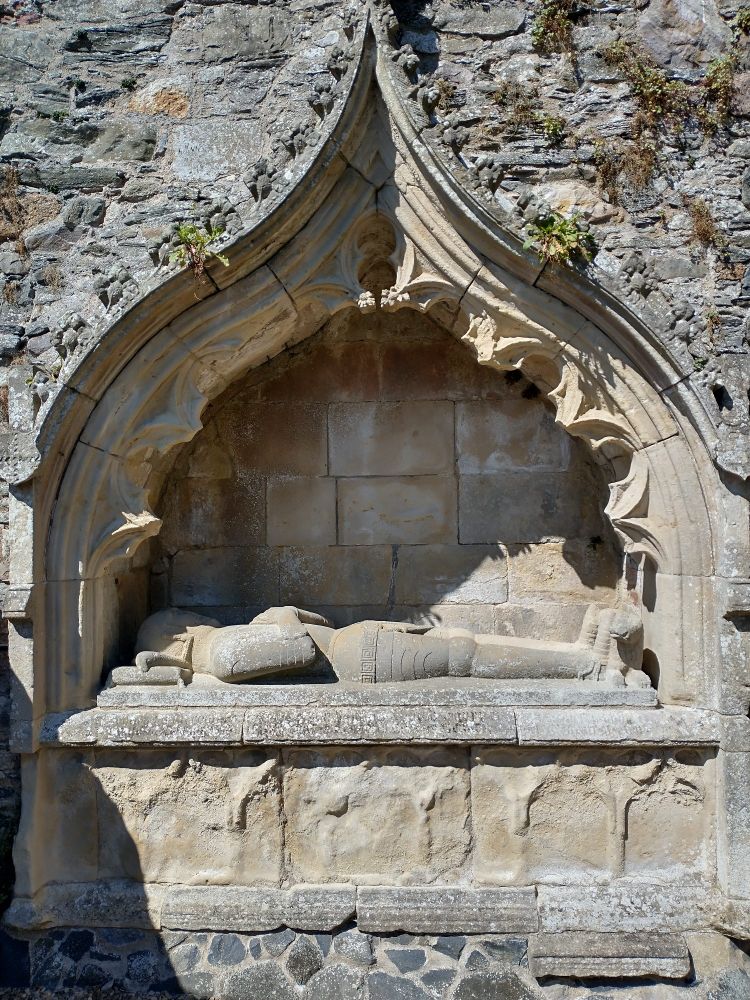 A carved stone effigy of a knight lies in a niche within a stone wall.
