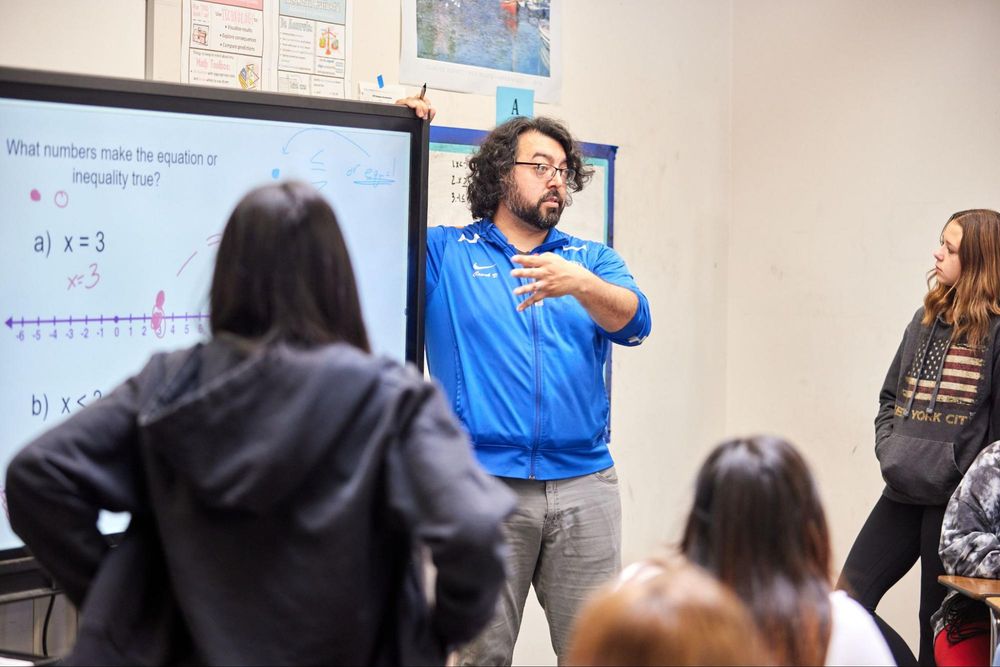 A teacher stands to a projector displaying classwork. A group of students stand around them and listen.