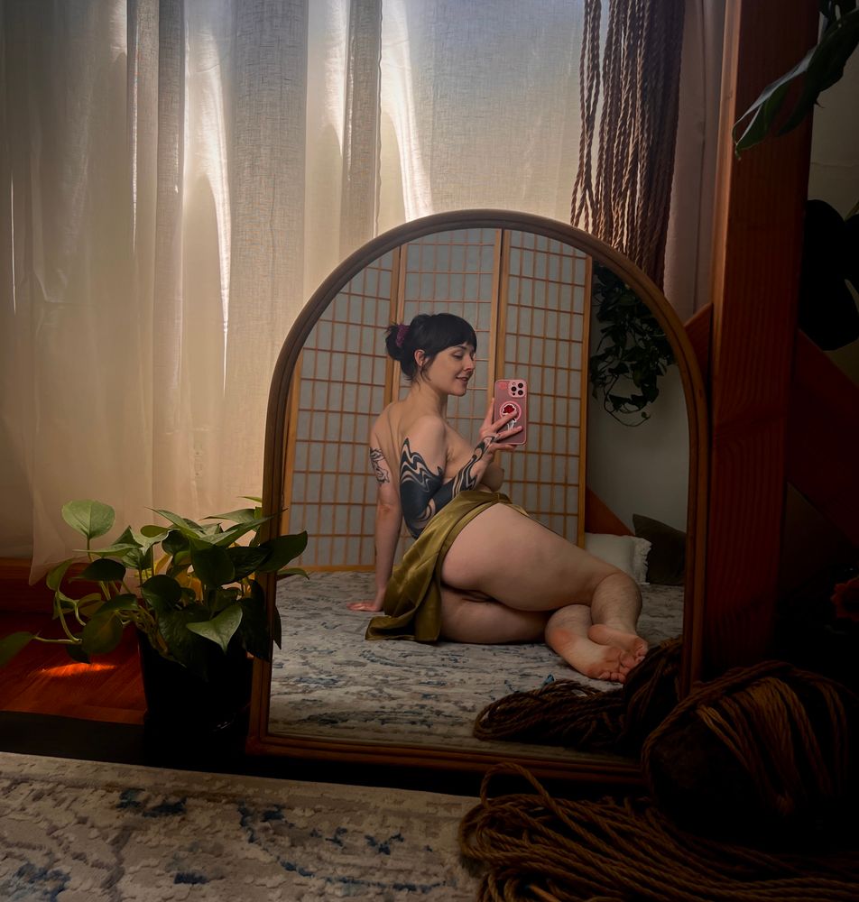 It’s golden hour, Ayala’s favorite time for photos. The light shines warm and bright through the thin linen window curtains. It’s a heavenly sight, especially when paired with the various plants in the room. Pythons, monstera, two name a few. Shibari ropes neatly bundled in their place, other rope is hanging up to stretch out and breathe. Ayala poses for a selfie in a small mirror. Their lift their olive green silk skirt up over their ass, revealing that there’s no underwear on them. You notice they’re also topless, you can see their nipple peeking ever so slightly with them sitting back facing the mirror but torso turned to face it. They show a little smirk at the camera. They have blackwork tattoos, their hair in a bun, and the cutest feet you’ve ever seen. 