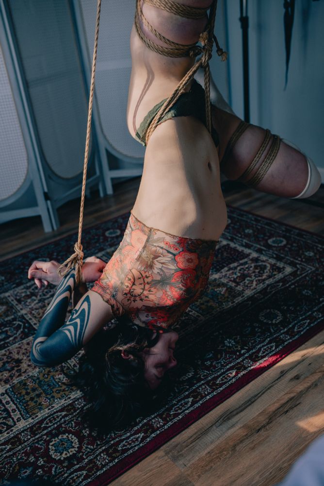 Ayala in a shibari rope suspension. Patches of sunlight enter the room