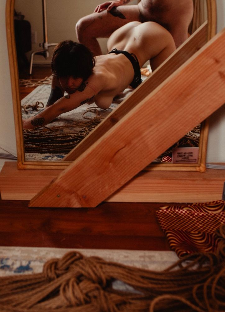 Camera is pointed towards a mirror, showing that Ayala is on their hands and knees in front of the mirror. Their arms are stretched out forward and back is arched as Knotsnacks is fucking them.