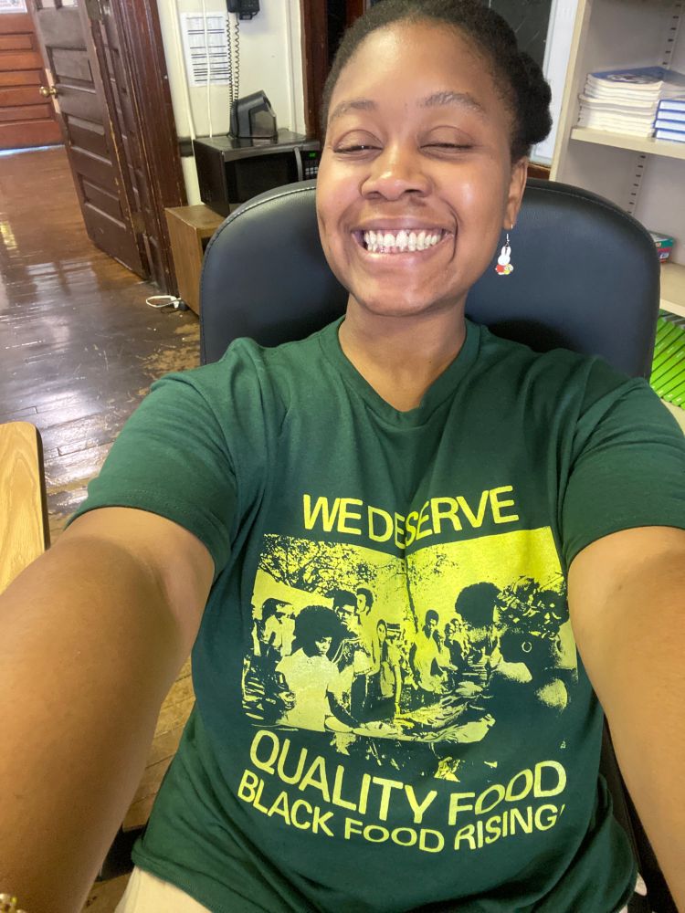 Richelle is a Black genderqueer person with Afro hair pinned back. They are smiling sitting in a black office chair showing a Black Food Shirt supporting co-op markets in Brooklyn, NY! 