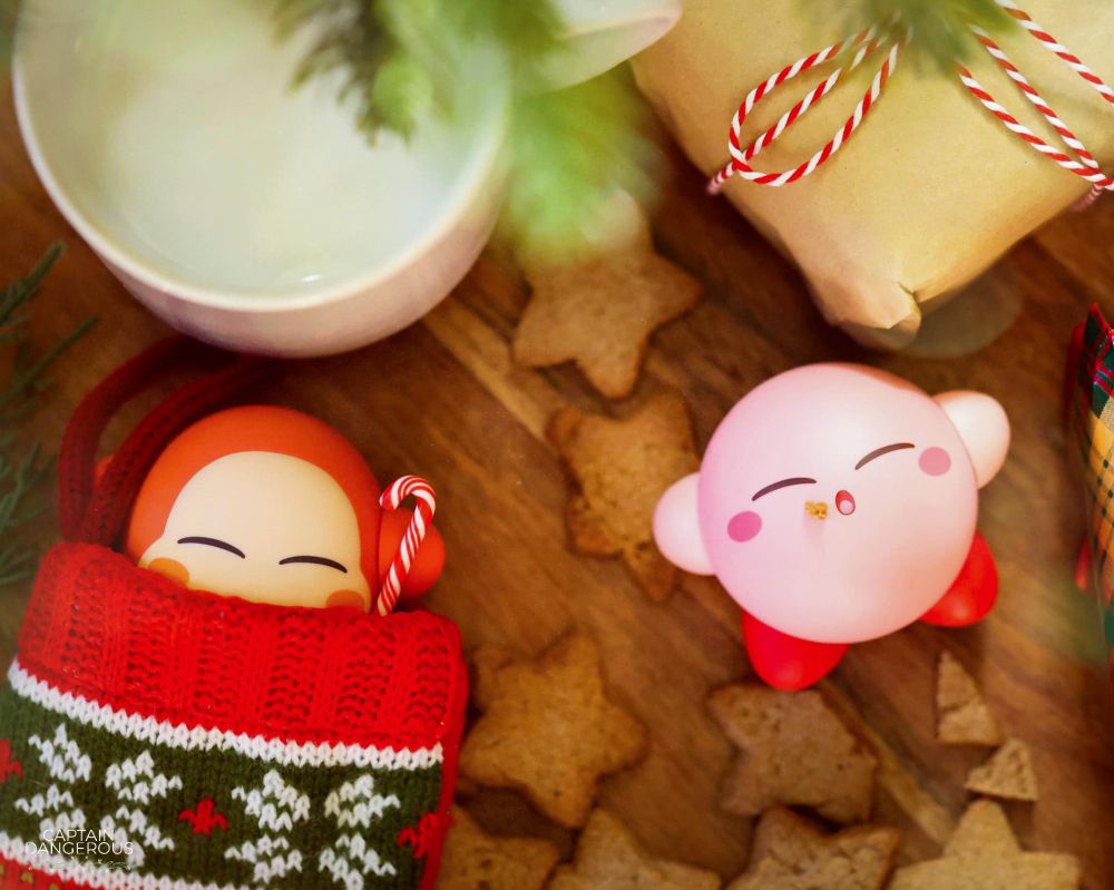 Miniature art of Kirby and Waddle Dee lying on a table among Christmas decor. Waddle Dee is partially in a stocking and holding a candy cane. They are surrounded by star-shaped Christmas cookies.