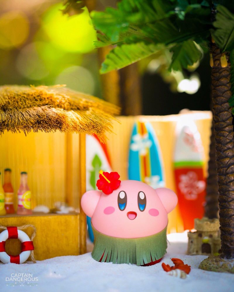 Miniature art of Kirby wearing a flower and grass skirt, standing in front of surfboards and by a small beachy outdoor bar counter.