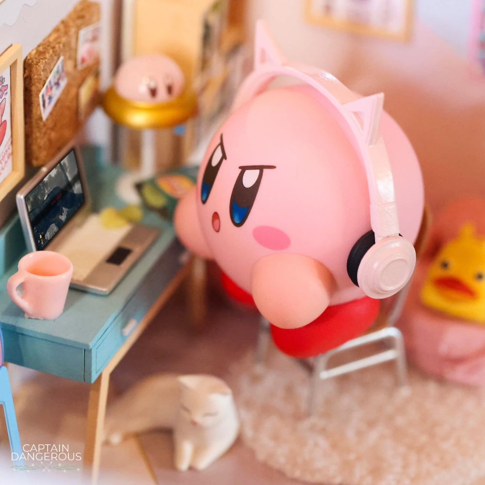 Miniature art by Captain Dangerous of Kirby playing Elden Ring on a laptop. He wears pink cat ear headphones and a cat sits beneath him.
