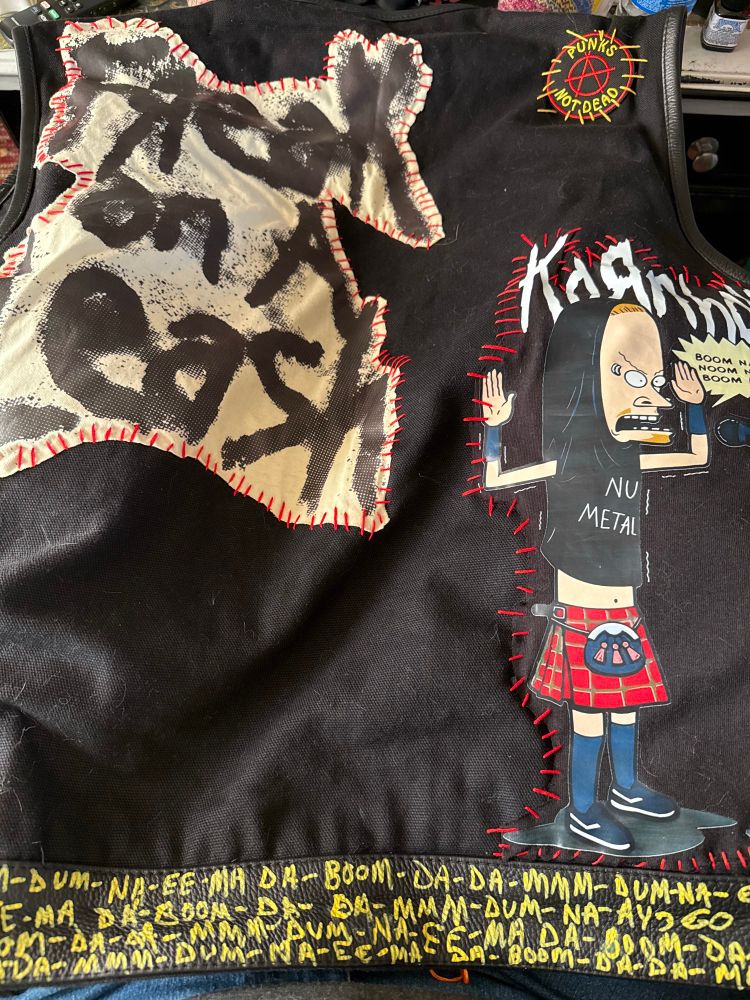 The beginning stages of a Korn vest. It is black with tons of pockets and on the leather strip along the bottom I included some of the lyrics to Freak On A Leash in yellow paint. Above that so far are 3 patches, 2 made from t-shirts. A Beavis and Butthead storyline of Kornholio with a black nu metal tshirt pulled over his head like a hood, a red plaid kilt with one of those kilt purses, blue stockings, black and white sneakers singing into a microphone with a stand of a modern art naked female. He is singing: Boom na da boom noom na nena boom na da boom. It is stitched in place with red embroidery floss, a circular Punks not dead with an anarchy symbol of an A encircled and its stitch is in yellow orange. Last patch so far is cream and black and says Freak on a Leash.