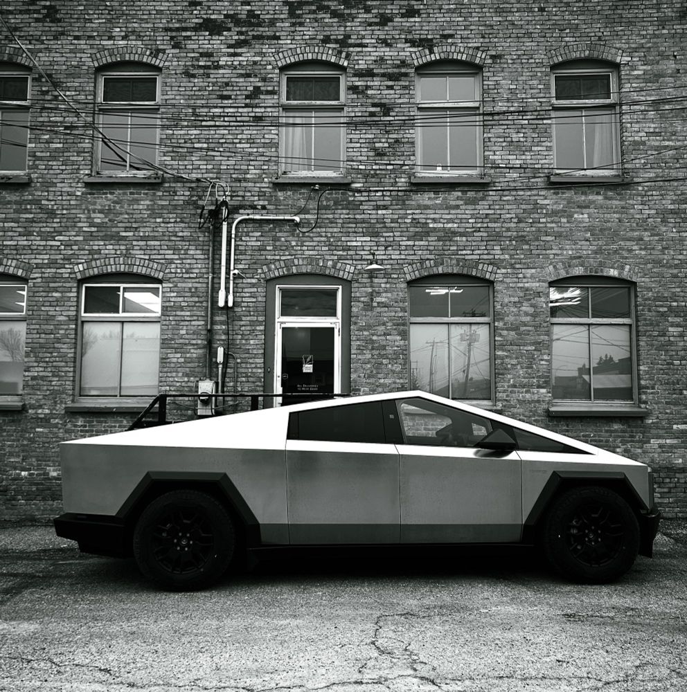 The image shows a futuristic, angular pickup truck parked beside an old brick industrial building. The truck has a geometric, stainless-steel–looking body with sharp edges, flat panels, and a wedge-shaped profile. Its wheels are large and dark, giving it a rugged stance.

The building behind it has a vintage, factory-style look: weathered brick walls, tall arched windows, and visible exterior conduit pipes running along the surface. The door at the center appears to be a simple commercial entrance with glass panes. The overall scene has a somewhat gritty, urban-industrial feel, and the black-and-white color tone enhances that mood.