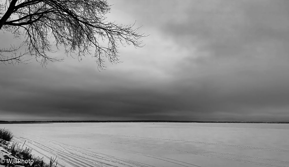 B&W image looking across a frozen over lake with a partial tree in the top right hand corner and a moody dark sky.