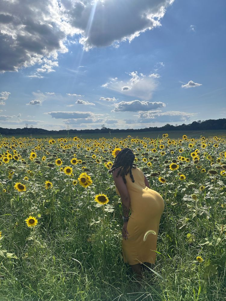 Black woman in a yellow dress in a sunflower field. She is standing with her back turned to the camera as she sniffs a sunflower. The sky has some cloud coverage with a fair amount of the sunrays shining through. 