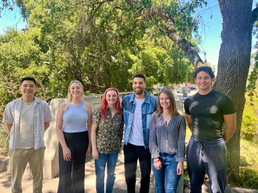Six young adults stand in a line outdoors, smiling in casual attire, with trees and sunlight in the background.
