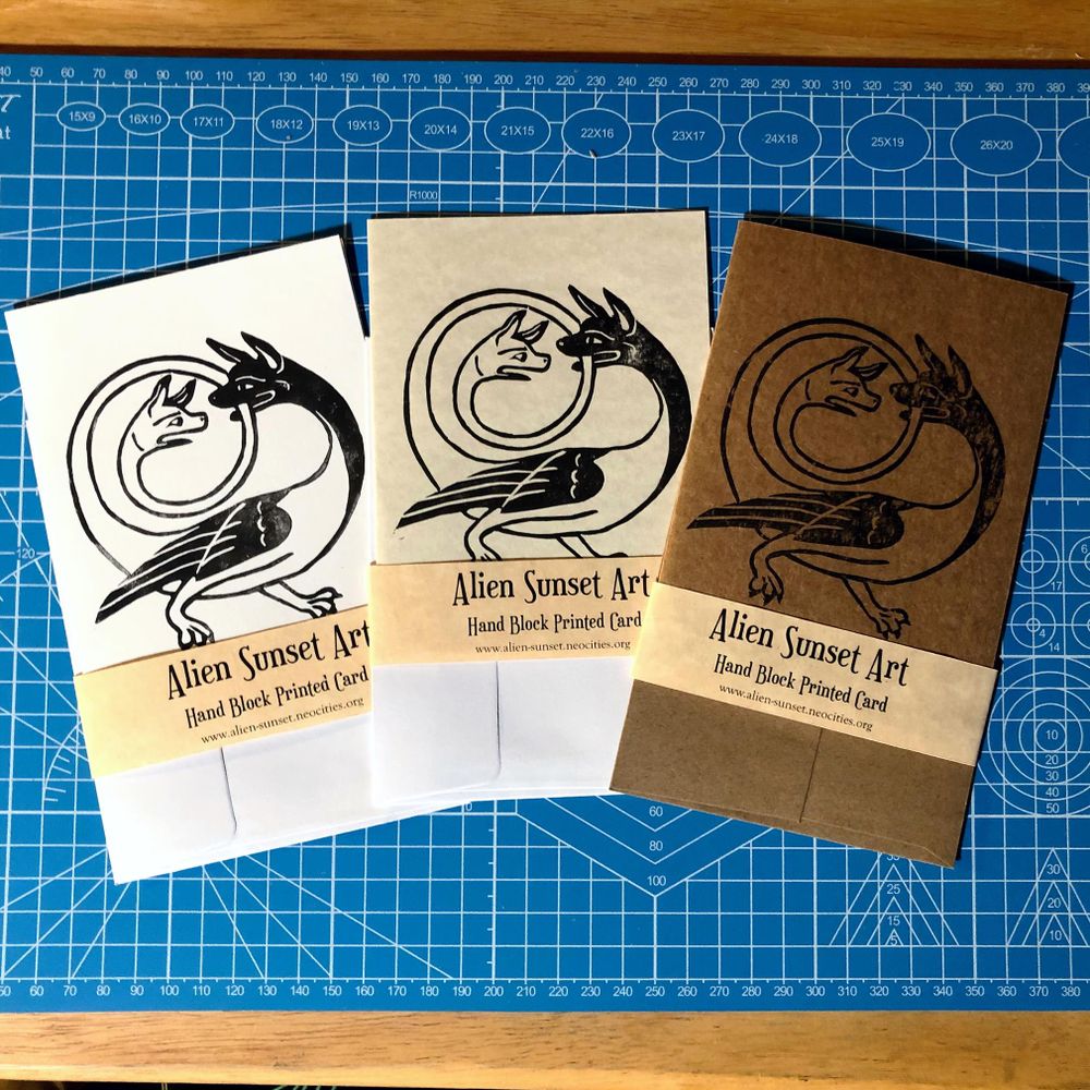 a set of three cards laid out on a blue cutting mat. a brown Kraft card, a plain white card, and a cream parchment pattered card. each card has a yellow parchment strip of paper wrapped around it that says "Alien Sunset Art" Hand Block Printed Card.

the block print on the cards is of  a creature with two heads, wings and two legs with paws, one head is situated at the end of a long tail that is forming a spiraling circle up and over the main body, the ’main’ Head is biting the tail as it circles near it and the second head is looking at it in shock/outrage/despair