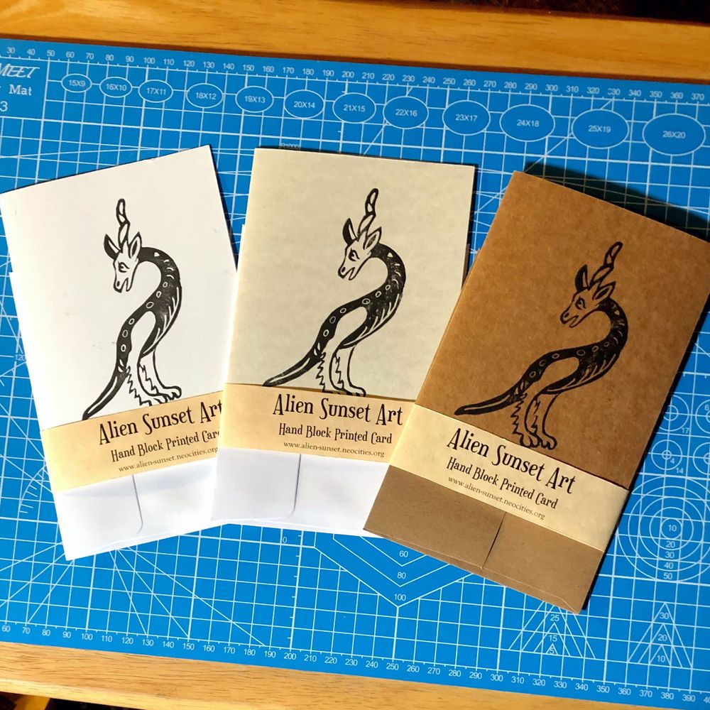 a set of three cards laid out on a blue cutting mat. a brown Kraft card, a plain white card, and a cream parchent pattered card. each card has a yellow parchemnt strip of paper wrapped around it that says "Alien Sunset Art" Hand Block Printed Card.

the block print on the cards is of a tall two legged creature with a long tail, furry legs and a horses head and unicorn horn