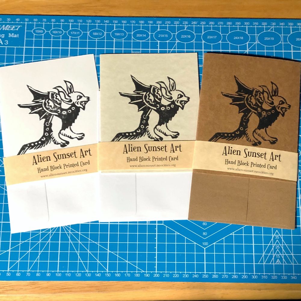 a set of three cards laid out on a blue cutting mat. a brown Kraft card, a plain white card, and a cream parchment pattered card. each card has a yellow parchment strip of paper wrapped around it that says "Alien Sunset Art" Hand Block Printed Card.

the block print on the cards is of  a small lizard creature with leathery wings and a devils face