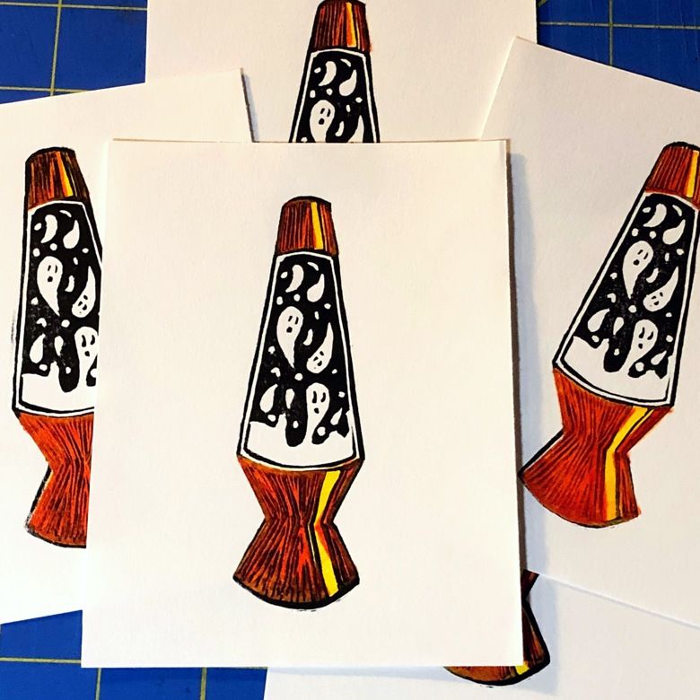 a photo of some block prints on white paper laid out on a table, the prints are of a lava lamp with an orange and black striped base and top cap with yellow highlights. Inside the main lamp, instead of the usual lava, there are white ghosts floating around on a black background in a similar manner.