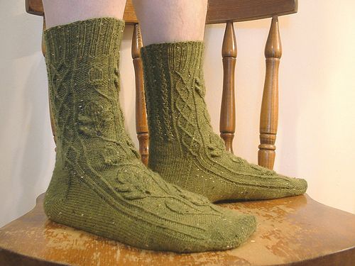 a pair of feet standing on a wooden kitchen chair, the feet are wearing a pair of lovely green socks with cables and oak leaf motifs.
