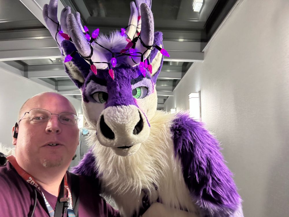 Me alongside a tall purple and white caribou fursuiter with green eyes that has purple lights strung through its antlers.