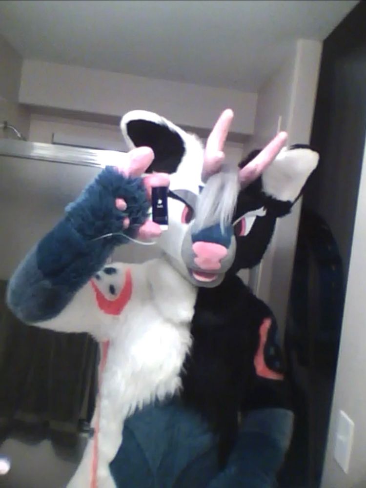 A fursuit of a white tailed deer holding an iPod nano 5th generation to take the photo in front of a mirror