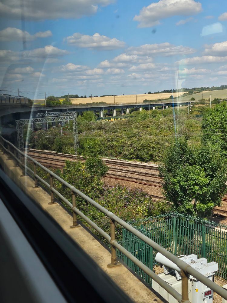 The view from a commuter train headed to Cambridge. The train is travelling along a raised bridge that curves and is visible ahead. On the right down below is train tracks and trees. Ahead is dry fields and blue sky with a scattering of clouds.