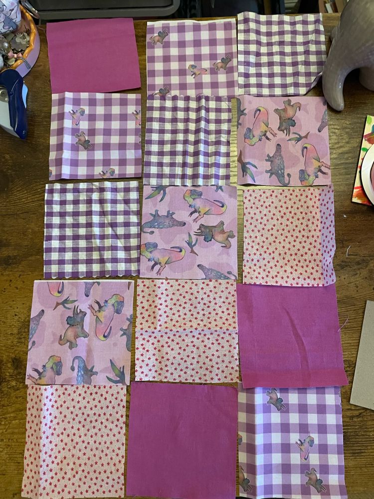 An overhead photo of a quilt layout. Square quilted pieces include: dinosaur fabrics, purple gingham and ditsy floral pink and red fabric