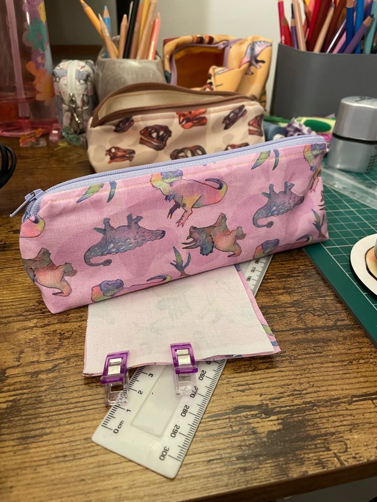 A photo of a purple and pink handmade pencil case, the fabric is also custom made and is decorated with tyrannosaurus, triceratops, pteranodon and ankylosaurus.