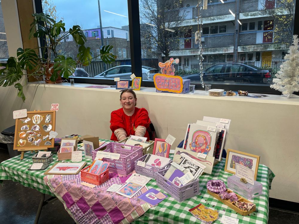 Me at my Queermas stall - green gingham table cloth with a pink and purple pastel gingham and dino-patterned quilt. Prints, stickers and hand-made scrunchies across the table.