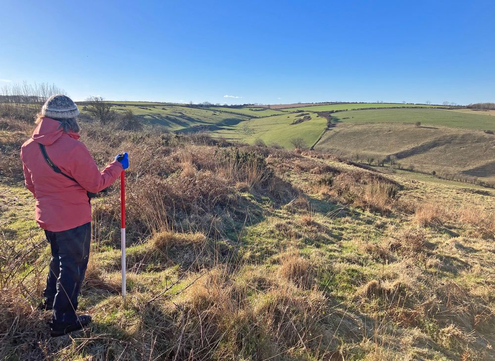 Colour photograph showing a person with their back to stading on moorland scrub looking out across a valley towards green pasture fields. The sky is bright blue and the far fields show remnants of prehistoric field banks within the modern plots. The person is wearing a pink coat, black trousers and a knited hat. They are holding a 1m red-and-white pole.