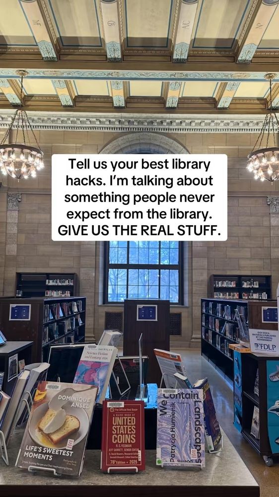 Interior of George Latimer Central Library featuring promotional book displays and banners, with a large caption that reads: 'Tell us your best library hacks. I’m talking about something people never expect from the library. GIVE US THE REAL STUFF.' The setting includes high ceilings, large windows, and classical architectural details.