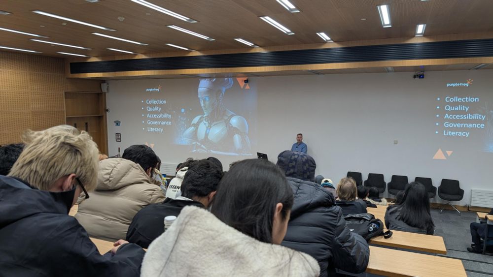 A lecture hall with tiered seating where attendees are listening to a presentation. At the front, Alex is presenting in front of a large screen displaying a slide with a futuristic robot graphic and a list of key terms: ‘Collection, Quality, Accessibility, Governance, Literacy.’ The room has wooden paneling, bright ceiling lights, and several people are seated with notebooks and laptops.