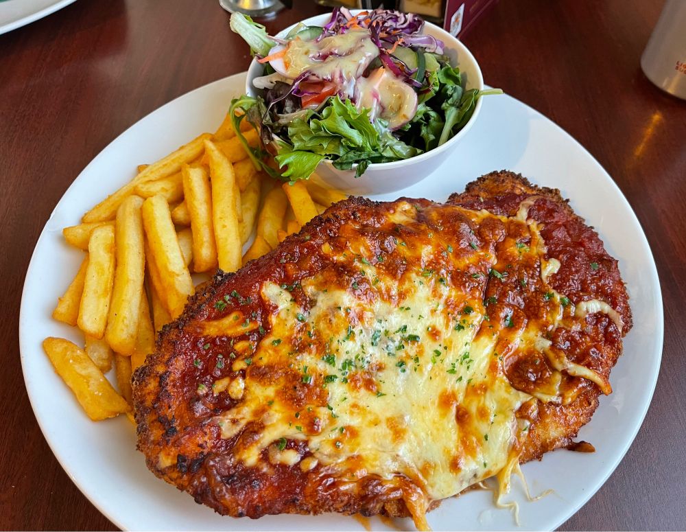 An enormous chicken parmigiana taking up 3/4 of a large plate, accompanied by chips and a small bowl of salad.
