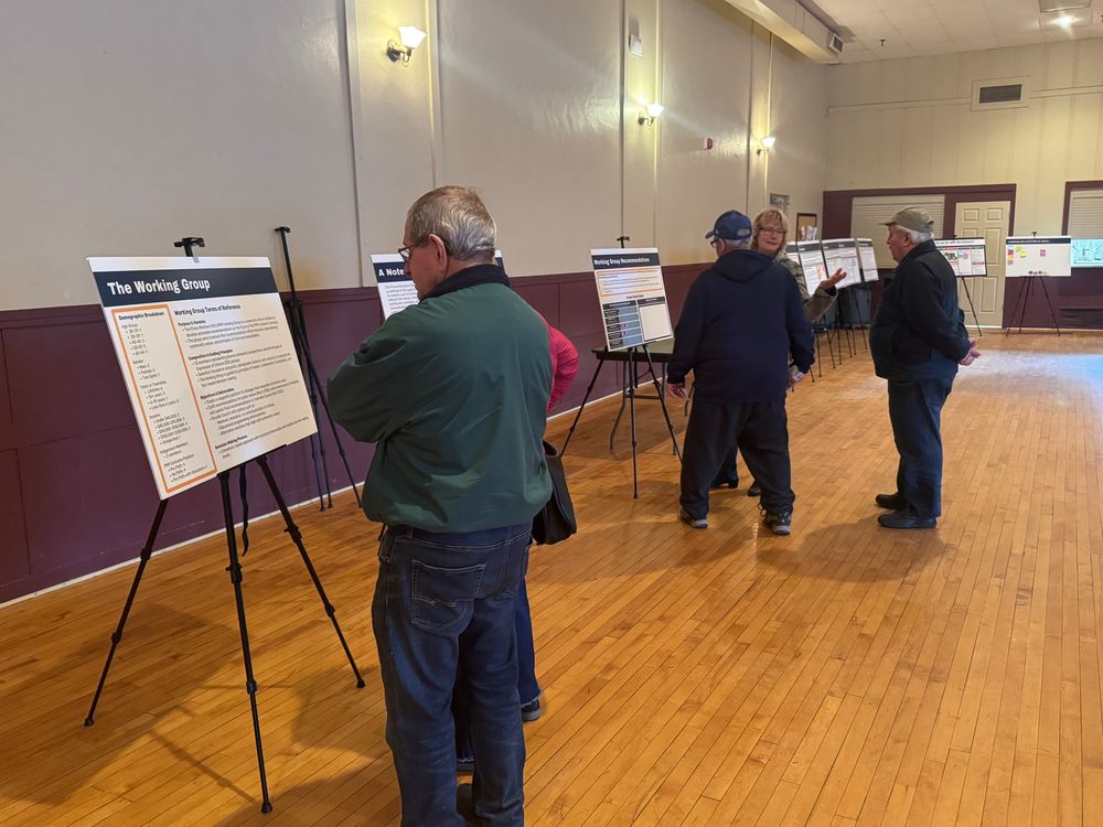  a picture in a room with white walls, purple wainscotting, and a narrow planked lightly stained wooden floor.  there are wall sconces with lights on the wall.  In the photo 5 people are congregated around a series of posters describing the Prime Ministers Path project and recommendations. 
