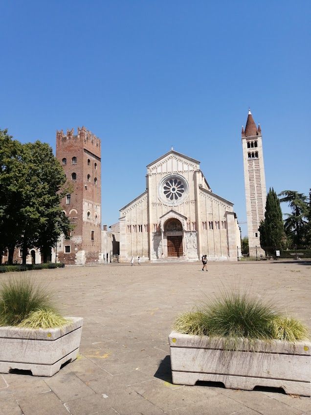A photo of the square in front of the Church of San Zeno in Verona. THe square is pedestrianised and a person is seen walking on it. The Church stands at the end of the square, with its steeple on the right, and a red-brick tower with merlons. The sky is blue and clear, it is a very sunny day. 