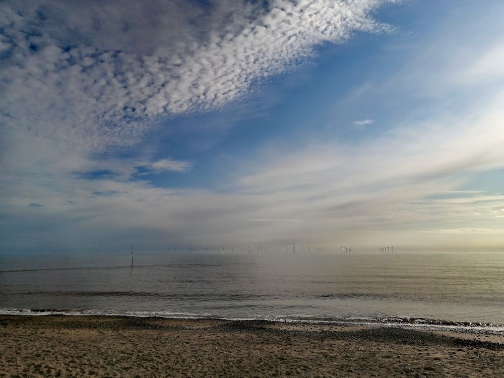 A view of the sea from a beach. The small waves are hitting the shore, and the sea is calm. In the distance we can see several small wind turbines. The sky is blue with some light white clouds. The light of the sun illuminates the scene from the right, giving some of the clouds a yellowish tinge, and is reflected in the water on the right-hand side. 