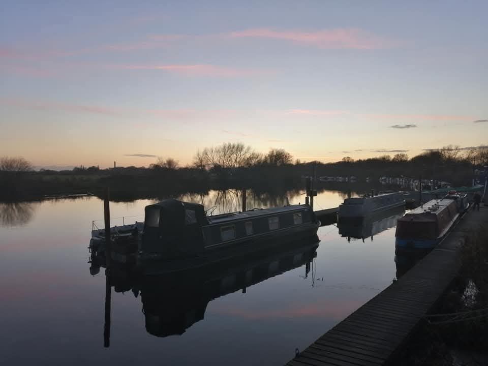 A calm river with a few sparse trees on the opposite side. It is afternoon and the sun is setting already. A few very light clouds have a soft rosy colour. Both the clouds and the trees are reflected in the clear water of the river. Closer to the viewer are some canal boats. The scene is very peaceful.