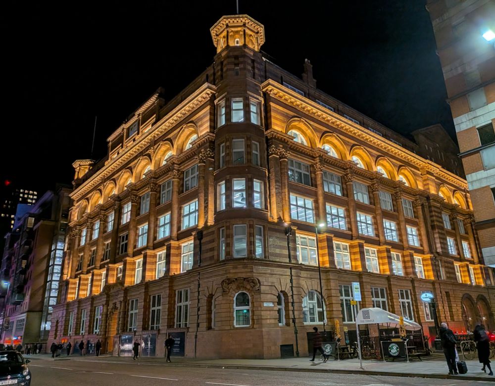 A large building with many windows and turrets of each corner, with red brick and white stone walls. It is dark and the building is beautifully illuminated.