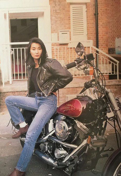 Maggie Chung in blue jeans, black shirt, and a black leather jacket, leaning against a parked motorcycle. my brain is running out of my ears and the goop is sparking on the ground