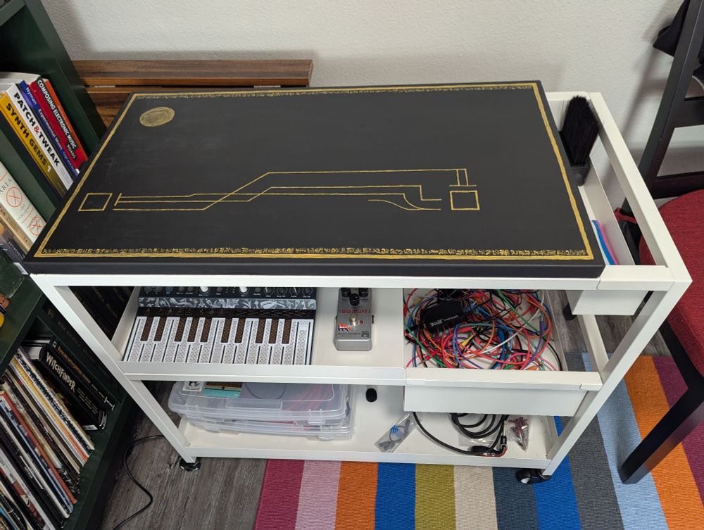 Ikea metal rolling cart with a bunch of music equipment + synths on it. The top surface has been painted black with gold designs 