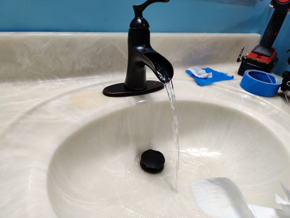 A picture of a sink with flowing water it is a dark oil rubbed bronze color in a faux marble basin. There are tools on the corner of the basin, a drill, painter's tape. A few partially out of frame. Water is flowing to show the sink works.