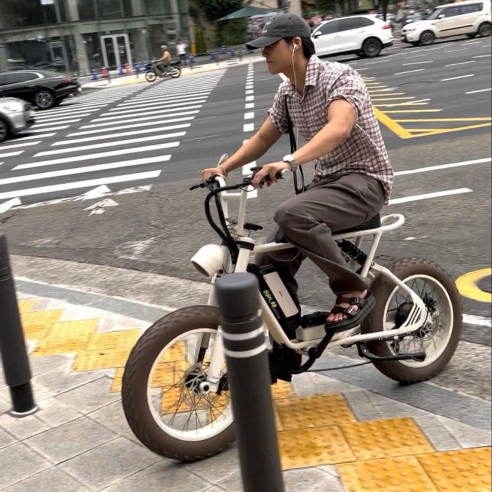 Kai andando de bicicleta, com boné, fones de ouvido, camisa xadrez e calça cinza