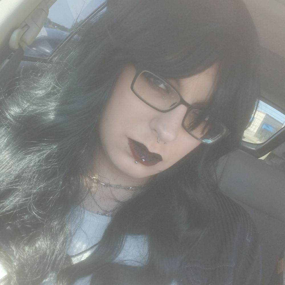 Selfie of rans. She has black and red ombre lipstick, and black hair. She is wearing a white crop top with a black overshirt and a silver necklace.