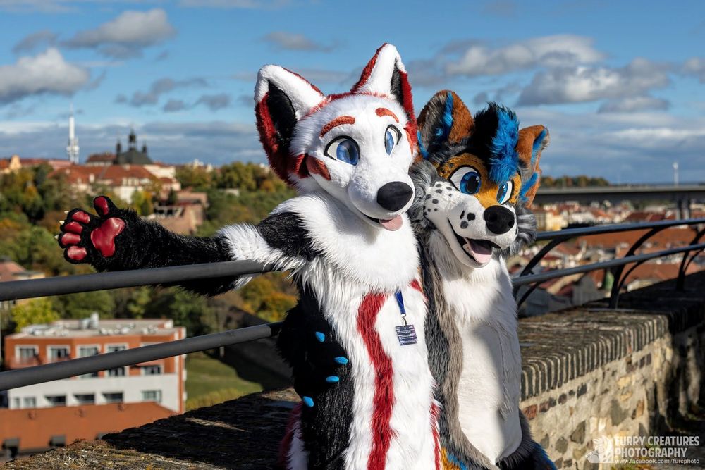 2 fursuiters leaning against a fence with a nice view of Prague behind them. The left fursuiter is a fox with white, red and black fur and the right one is a shepherd with grey, white, brown and blue fur. Both look very happy with opening arms!
