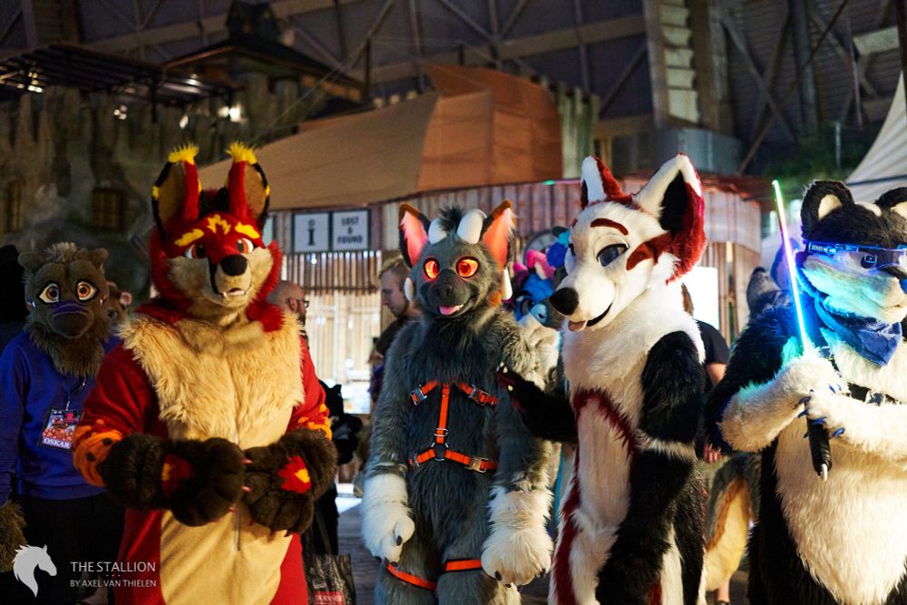 5 fursuiters, standing in a decorated aquapark. From left to right, there is a brown otter with purple markings, a red, beige and yellow fox, a gray and white goat, a white and black fox with red markings and a black and white fox. The last one has a light saber in its paw.