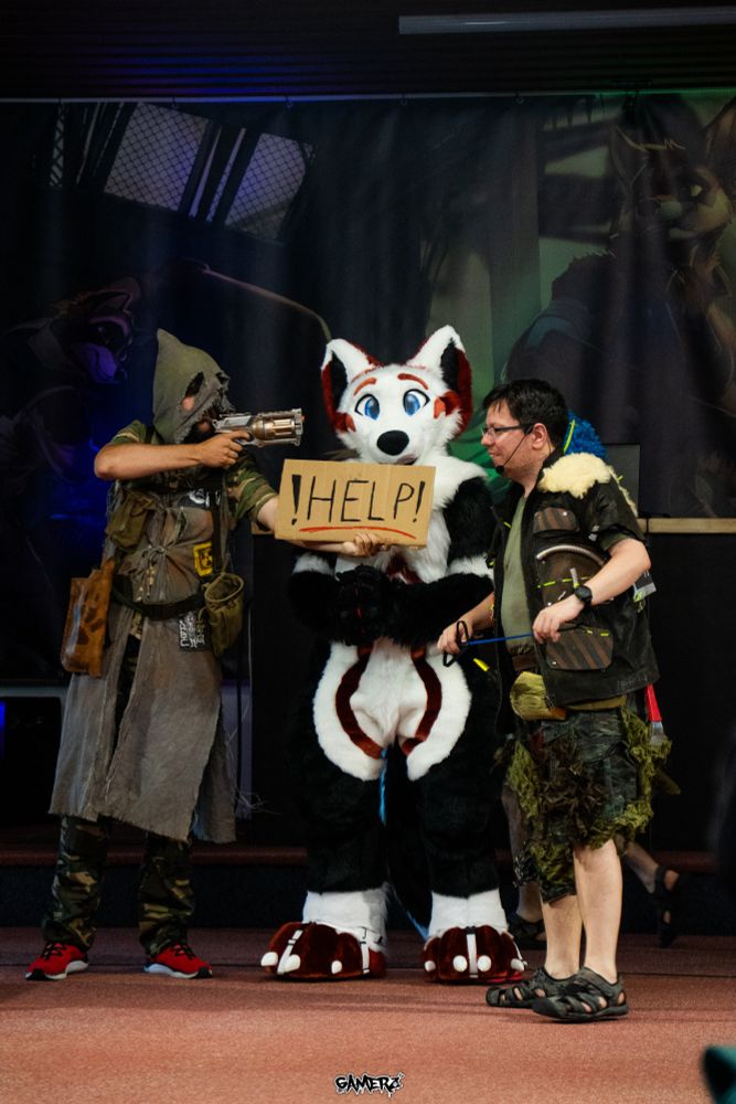 A white, red and black fox fursuiter standing inbetween two wasteland dressed cosplayers. The fursuiter is holding a sign "HELP", while the right cosplayer is smiling in an evil way and the left cosplayer is holding a gun pointed at the fursuiters face. 