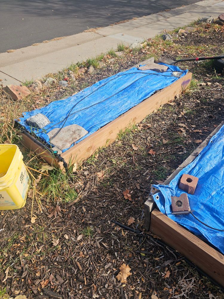 Slightly zoomed out wooden garden boxes, covered only in the dirt areas, with blue tarps weighted down by rocks in the corners. 