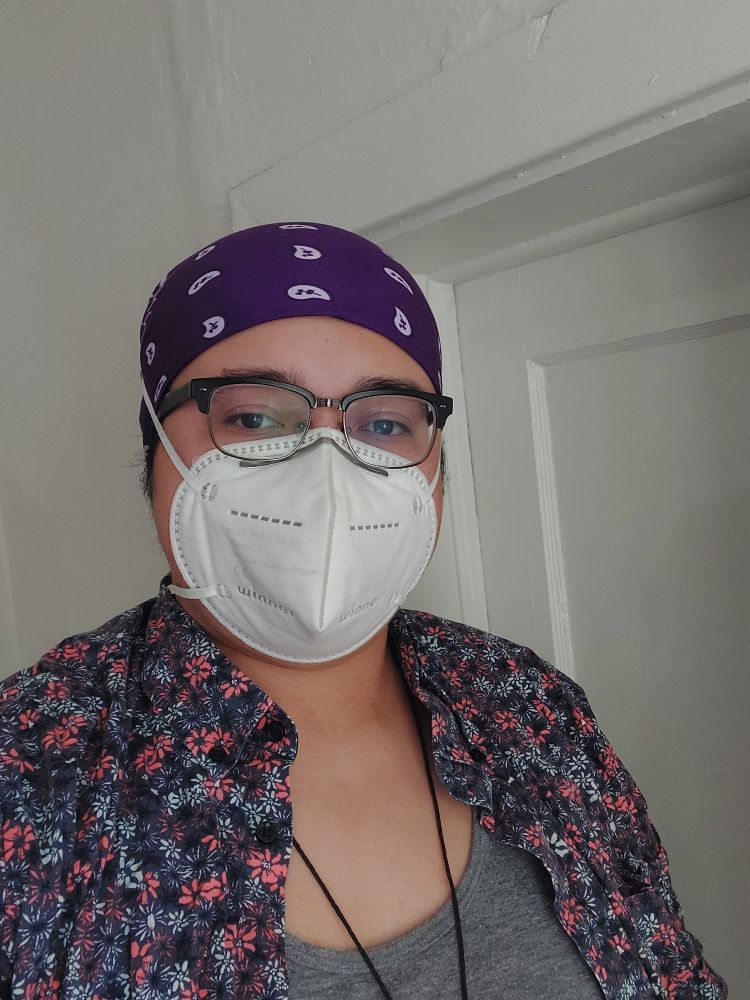Image is a selfie of a brown-skinned Latino person, wearing a purple bandana on their head, black glasses and a white KN-95 mask. As well an open purple and orange floral print button-down and a gray undershirt