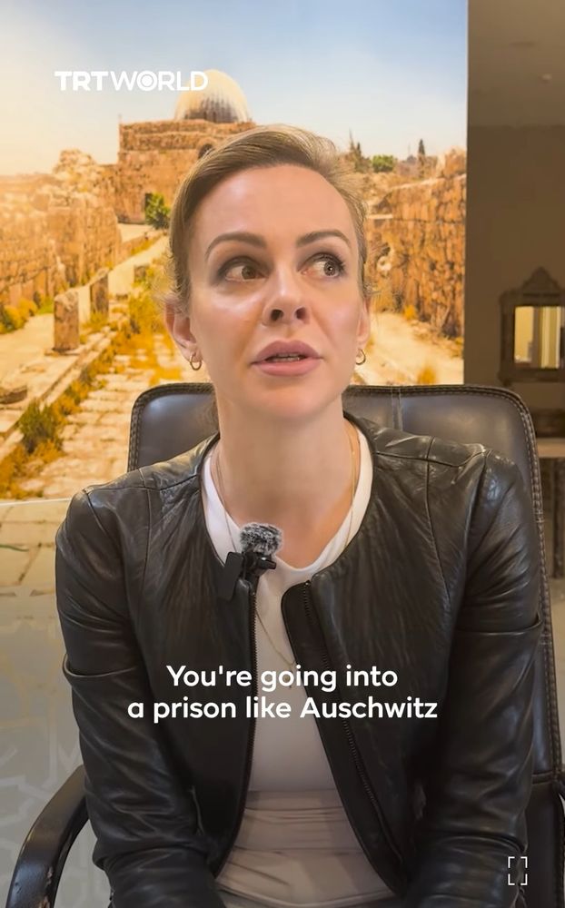 Screenshot from linked vid: a white woman wearing a white tee & a black leather jacket, open caption reading "You're going to a prison like Auschwitz."