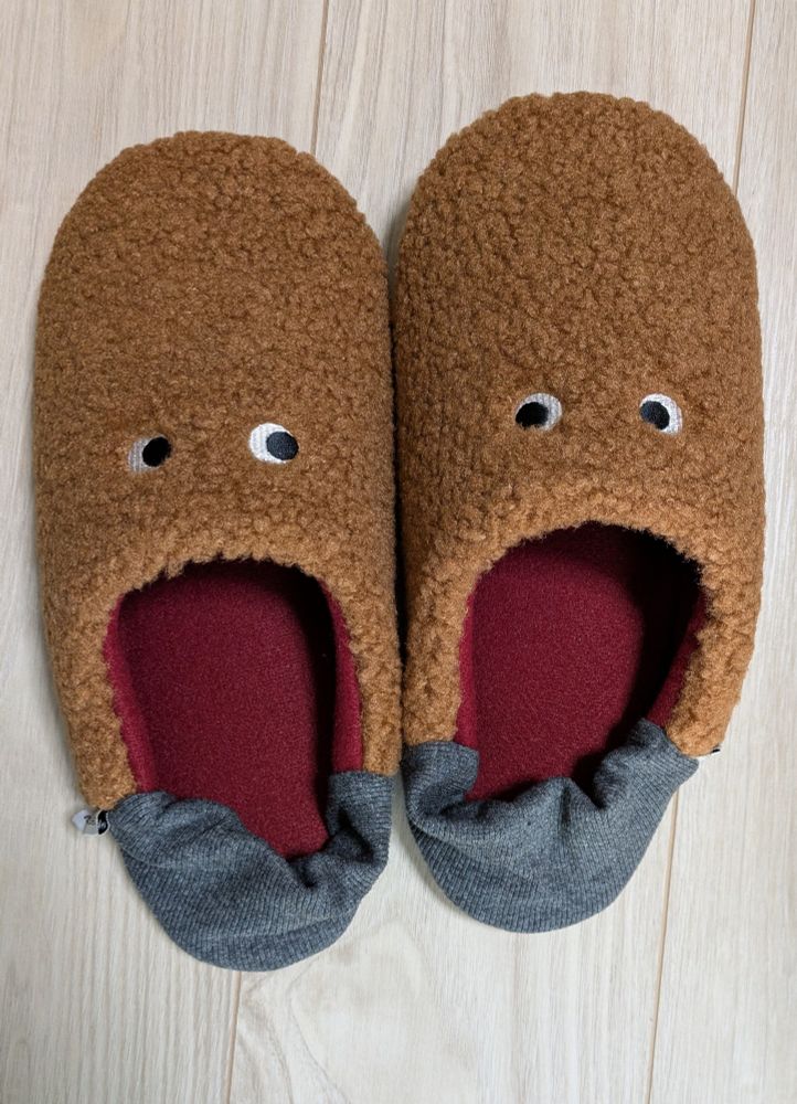 新しく買ったルームシューズの写真。足の甲部分が茶色いモコモコで、キョロ目の形の刺繍がしてある。加えて中敷きが赤なので、脱いで置いてあるとモコモコのお化けが叫んでいるように見えるデザイン。