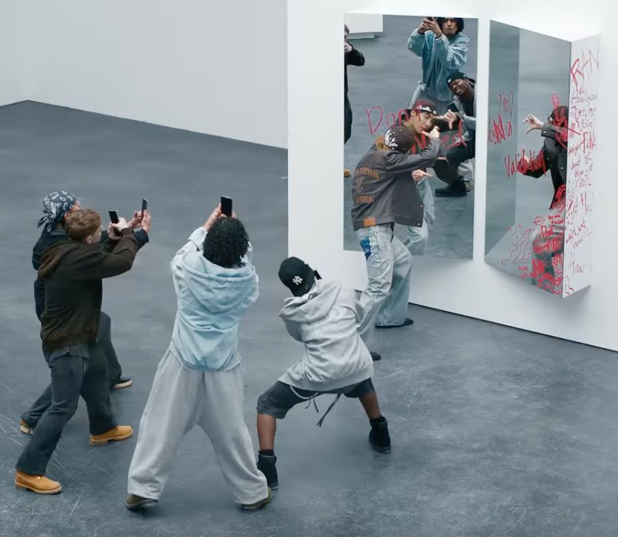 Screenshot from Jhope's "Mona Lisa" MV where he is looking in a mirror with writing on it. Grey background and floors. His background dancers are pretending to take photos of him on their phones.