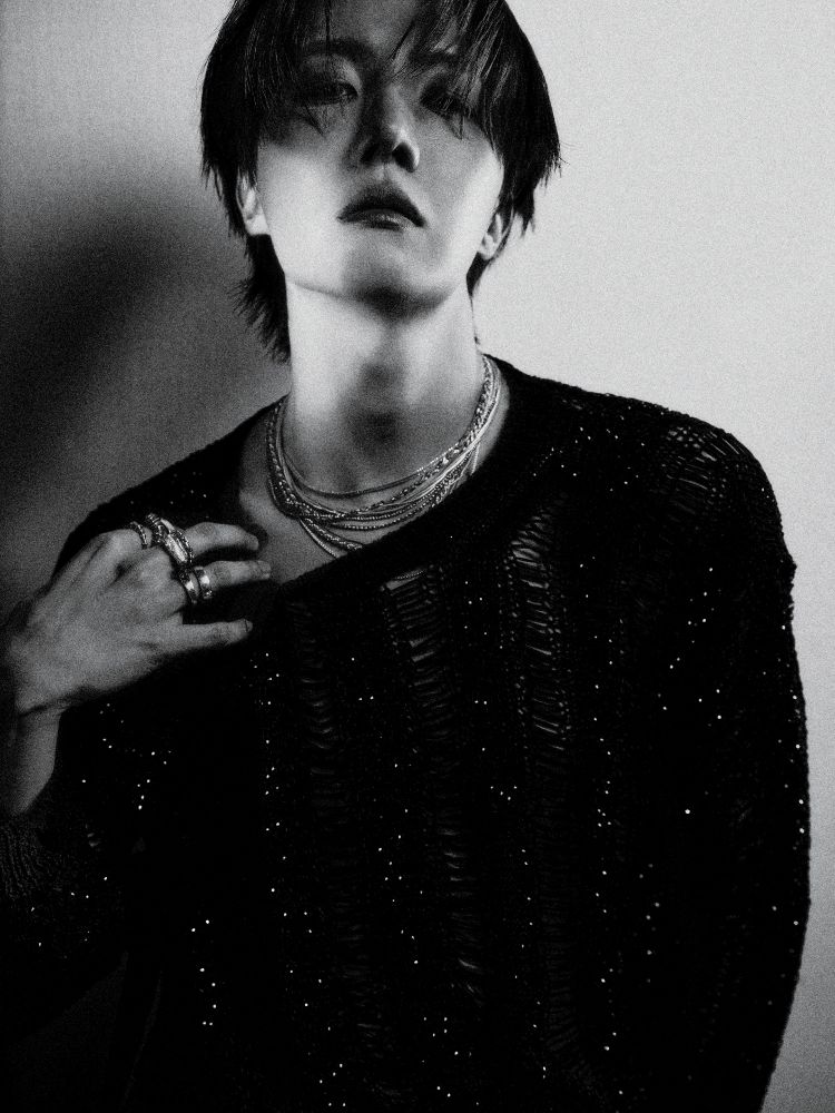 B&W of Jhope concept photos. He's staring into the camera, chin up, wearing a loosely stitched sweater, necklaces, and rings. He is pulling down one side of the sweater's neck in a clear invitation to notice his lack of mating bite...