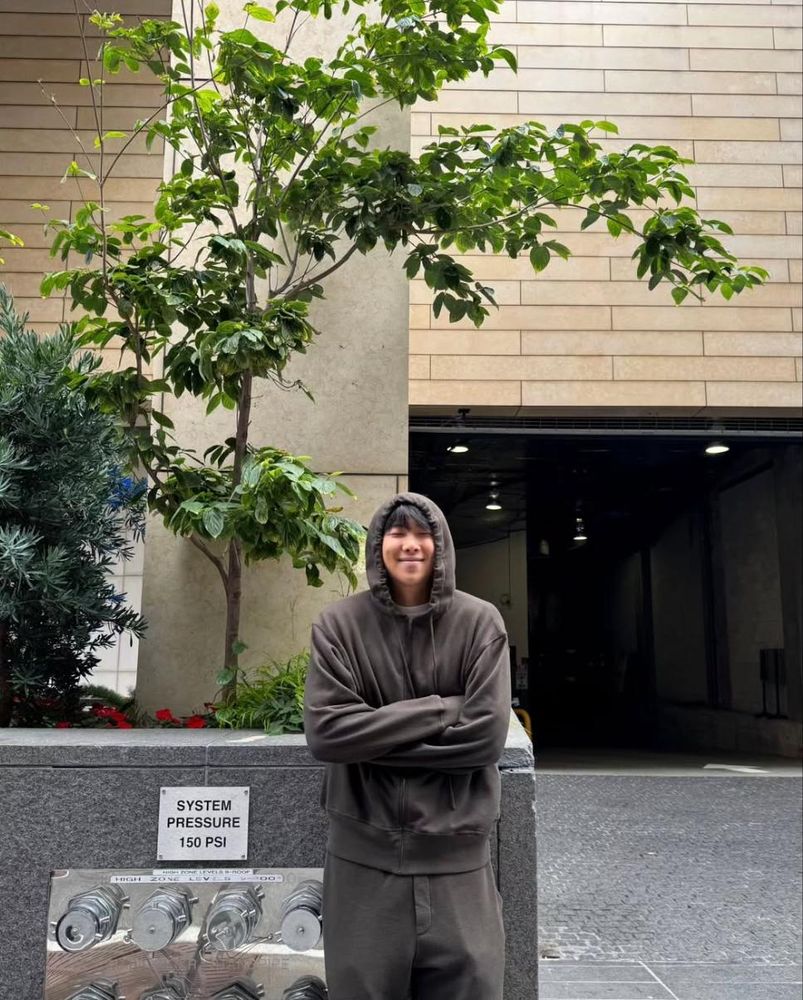 Namjoon, grinning with his arms crossed in a brown hoodie, standing under a young tree and next to a sign that says "system pressure" that is above some pressure valves. He's standing outside a parking garage. From his Insta.