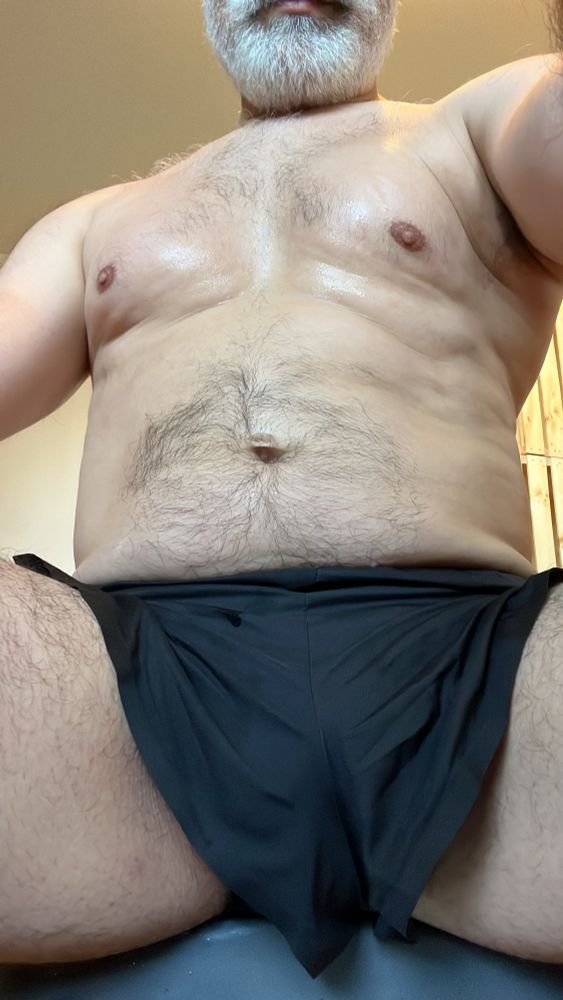 A picture of Kameron’s body after a sweaty 30 minute rowing workout at home. The picture is taken looking up between his spread legs, you can see his smile and grey beard, and the only piece of clothing seen is a pair of black Nike split-side running shorts. The blue swim-style jockstrap he is wearing underneath cannot be seen but its supportive nature can be seen in the bulge in his shorts.